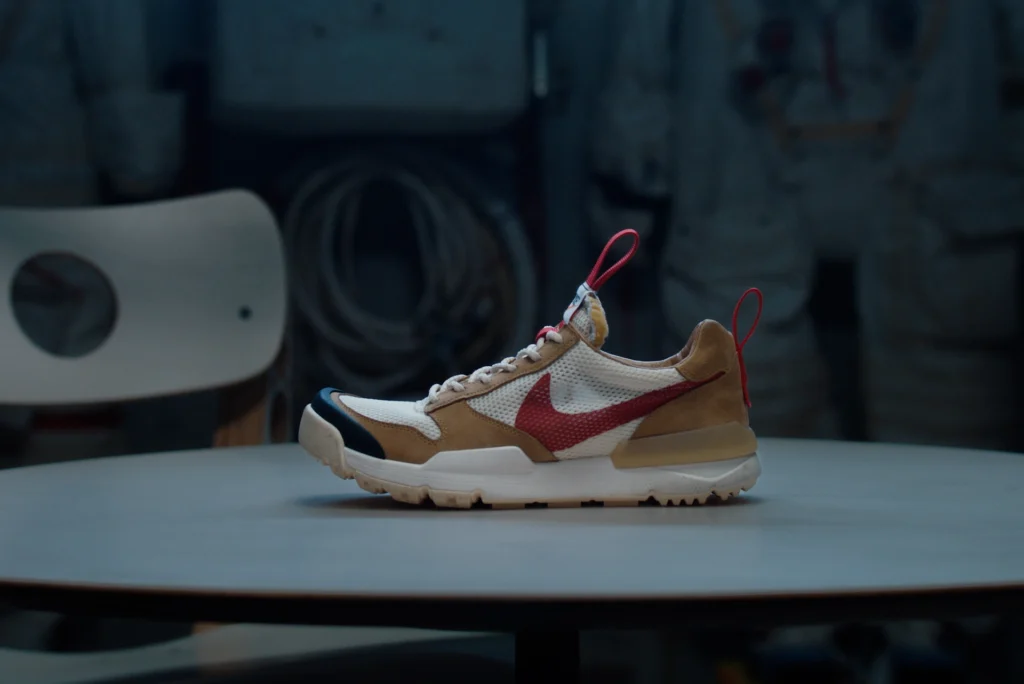 Side profile of the Tom Sachs x NikeCraft Mars Yard 3.0 sneaker, featuring a tan suede and white mesh upper with a bold red Swoosh, black rubber toe cap, and red pull tabs, displayed on a table with astronaut gear blurred in the background.