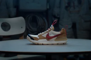 Side profile of the Tom Sachs x NikeCraft Mars Yard 3.0 sneaker, featuring a tan suede and white mesh upper with a bold red Swoosh, black rubber toe cap, and red pull tabs, displayed on a table with astronaut gear blurred in the background.