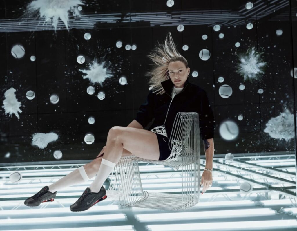 Belarusian tennis player Aryna Sabalenka wearing Nike Shox Z sneakers in a futuristic campaign setting, seated on a wire chair with dynamic background visuals.