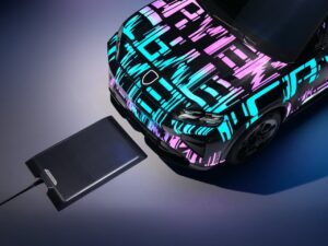 Prototype Porsche Cayenne EV parked over a wireless charging pad, featuring neon geometric camouflage pattern, illustrating Porsche’s inductive charging technology debuting in 2026.
