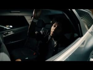 YoungBoy Never Broke Again sits in the backseat of a car at night, raising his hand toward the open window, wearing a black jacket and jewelry.