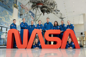 NASA’s 2025 astronaut candidates pose in blue flight suits behind a large red NASA logo inside Johnson Space Center, marking the agency’s first majority-women class.