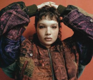 Lola Young poses against a burnt-red backdrop wearing a multi-tone denim and corduroy patchwork jacket, her hands resting behind her head, evoking the raw, introspective energy.