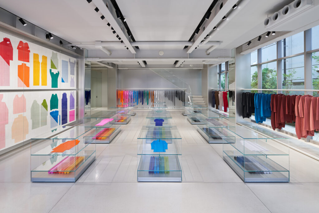 Interior view of the PLEATS PLEASE ISSEY MIYAKE flagship store in Aoyama, Tokyo, designed by Tokujin Yoshioka. The minimalist space features glass display tables with neatly folded pleated garments in vibrant colors, wall-mounted textile art, and natural light streaming through large windows, highlighting the collection’s signature interplay of color, structure, and light.