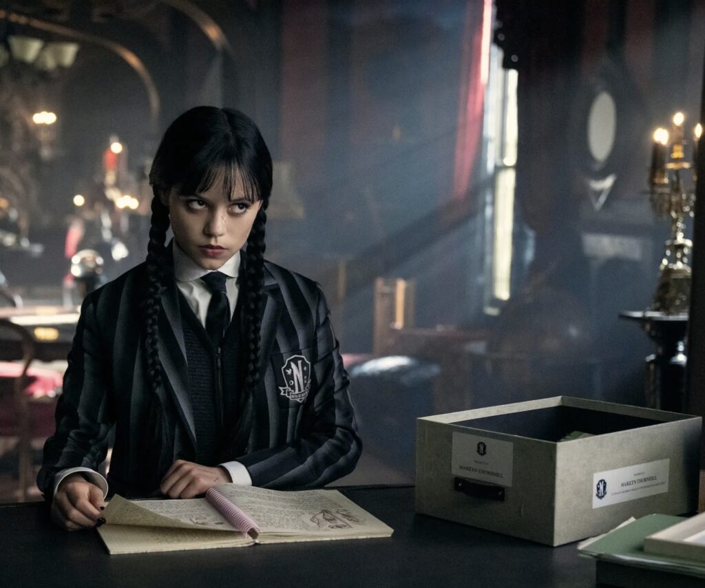 Jenna Ortega as Wednesday Addams in Netflix’s “Wednesday,” wearing a black-and-grey striped blazer with braided hair, sitting at a desk reading an open notebook in a dimly lit gothic room.