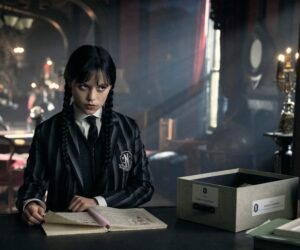 Jenna Ortega as Wednesday Addams in Netflix’s “Wednesday,” wearing a black-and-grey striped blazer with braided hair, sitting at a desk reading an open notebook in a dimly lit gothic room.