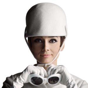 Portrait of Audrey Hepburn wearing a sculptural white helmet-style hat and white gloves, holding a pair of round white sunglasses against her chin, photographed in 1965 by Douglas Kirkland against a plain white background. The image highlights her poised expression, minimalist fashion, and the refined mod aesthetic of the mid-1960s.
