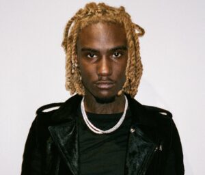 OsamaSon wearing a black jacket and layered silver chains, standing against a plain white background.