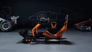 Side profile of the PLAYSEAT® Formula Instinct McLaren Racing Edition simulator, featuring an aerodynamic papaya-orange frame with integrated racing seat, steering wheel mount, and pedal base, set against a dark backdrop with Formula 1 car outlines and racing tires.