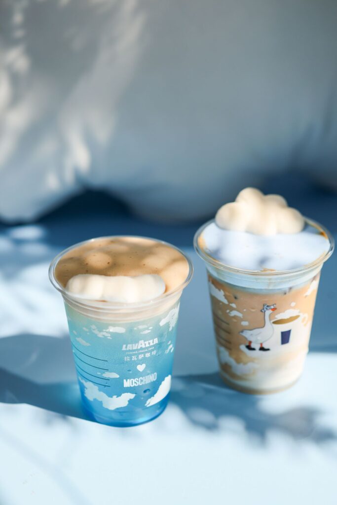 Two iced coffee drinks from the Lavazza × Moschino “Espresso Your Style” collaboration, served in limited-edition cups decorated with cloud motifs and a playful goose illustration, each topped with fluffy, cloud-shaped foam against a soft blue background.