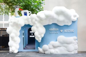 The entrance of the Lavazza × Moschino “Espresso Your Style” pop-up on Anfu Road in Shanghai, featuring a whimsical cloud-shaped installation in white and blue with a large Lavazza cup and a paper-hat-wearing goose perched above, surrounded by lush green trees.