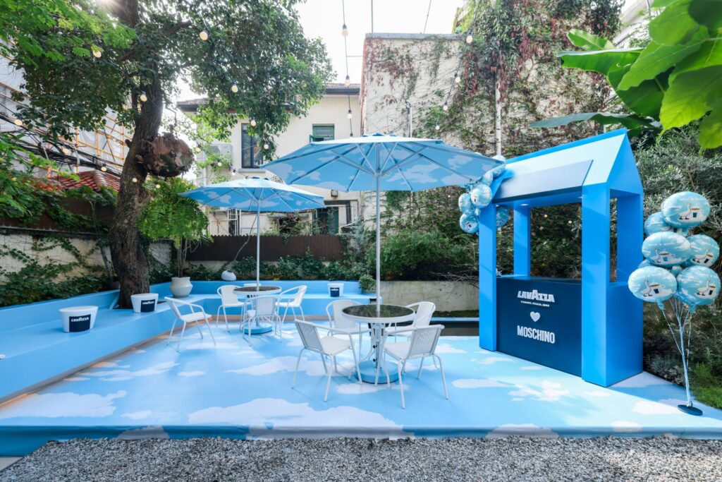 The outdoor Cloud Garden Terrace of the Lavazza × Moschino “Espresso Your Style” pop-up in Shanghai, featuring sky-blue flooring with white cloud patterns, round tables under matching parasols, string lights in the trees, and a Lavazza-branded booth accented with Moschino-themed balloons.