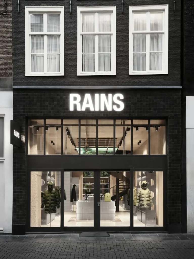 Exterior view of the RAINS store in Amsterdam showcasing the brand’s minimalist design language, with a black brick façade, large front windows displaying neutral-toned puffer jackets, and a glowing RAINS logo above the entrance.