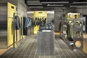 Interior view of the RAINS retail space featuring steel fixtures, modular shelving, and yellow accent walls, displaying minimalist outerwear and bags arranged on industrial racks under linear ceiling lights.