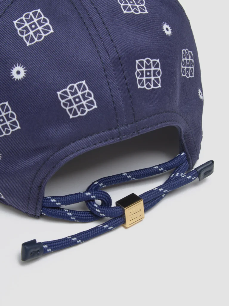 Close-up rear view of the Miu Miu Printed Cotton Baseball Cap in blue and white, showcasing the adjustable navy cord with white stitching detail, a gold-tone square toggle engraved with the Miu Miu logo, and the patterned cotton fabric with white geometric motifs.