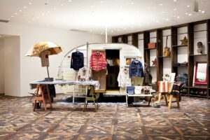 Interior view of an Indigo Camping Trailer pop-up installation featuring a vintage white camper surrounded by Visvim garments. A checkered table displays shirts and jackets dyed with indigo and mud, while shelves in the background hold guitars, textiles, and rustic décor, reflecting the brand’s fusion of craftsmanship, travel, and natural materials.