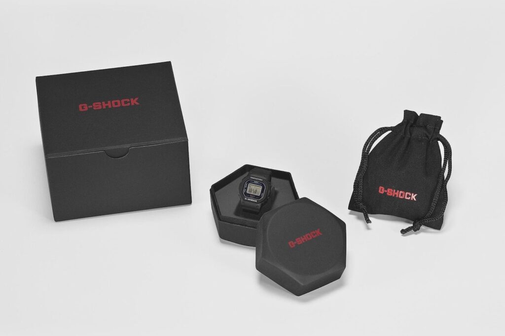 Product display of the G-SHOCK Nano DWN-5600 ring presented in its official packaging. The image shows a matte black box with red G-SHOCK lettering, an open hexagonal case containing the miniature digital ring watch, and a drawstring pouch with matching branding, all arranged on a clean white surface.