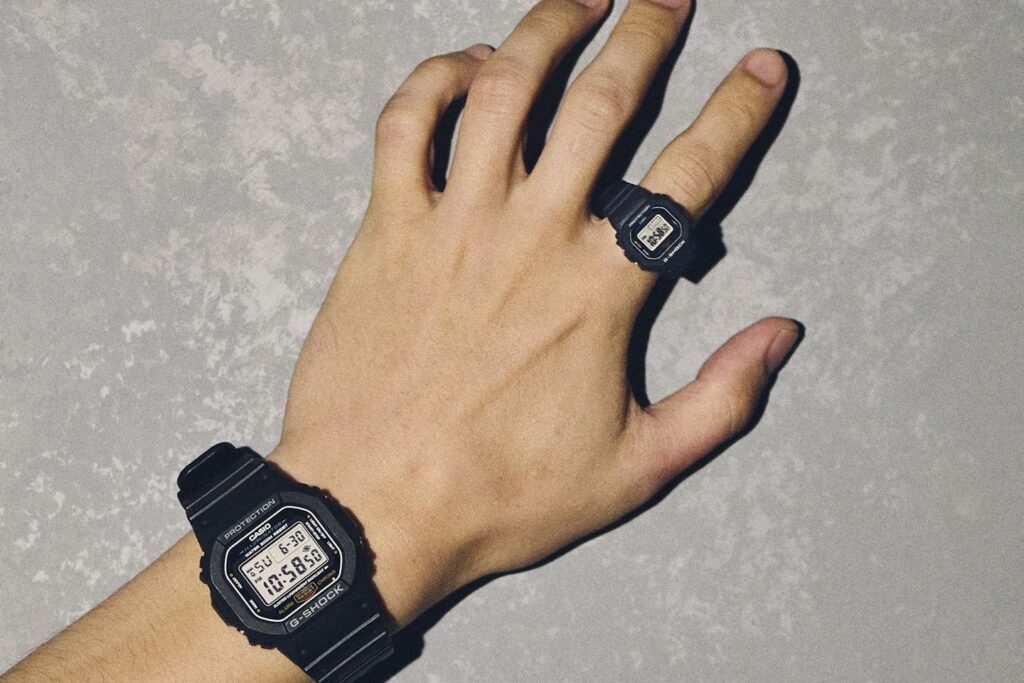 Close-up of a person’s hand wearing both a full-sized black G-SHOCK DW-5600 watch on the wrist and the new miniature G-SHOCK Nano DWN-5600 ring on the middle finger. The image emphasizes the size contrast between the original model and the ring-sized version against a textured gray wall background.