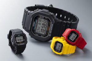 Four Casio G-SHOCK digital timepieces displayed together, showing a size comparison between the full-sized DW-5600 model and the new miniature Nano DWN-5600 rings. The collection features matte resin finishes in black, red, and yellow, each with the signature square bezel and “PROTECTION” inscription, placed on a neutral gray background to emphasize the contrast between the standard watch and the ring-sized variants.