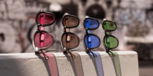Four pairs of MOSCOT Lemtosh sunglasses displayed side by side on a concrete ledge, each featuring the brand’s signature black acetate frames with vibrant transitional Tint+ lenses in rose, amber, blue, and green. Sunlight highlights the gradient hues, casting colored reflections on the surface below against a softly blurred urban background.