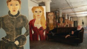 Francesco Clemente reclining on a dark velvet sofa inside his studio, surrounded by large, expressive portrait paintings of women with oversized eyes and solemn expressions, arranged against tall columns in a dim, industrial space.