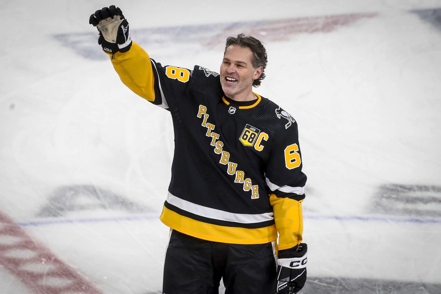 Jaromír Jágr smiles and raises his arm to greet fans while wearing a black and yellow Pittsburgh Penguins jersey with his iconic number 68 and captain’s “C.” He stands on the ice rink, celebrating during a tribute event, symbolizing his long and legendary hockey career.