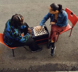 Two people sit on red chairs outdoors playing chess. The woman, wearing a denim jacket, red skirt, and black PUMA Suede sneakers, reaches to move a piece, while the man in a patterned blue-and-black sweater observes. The scene highlights casual street style and the Ahluwalia x PUMA collaboration.