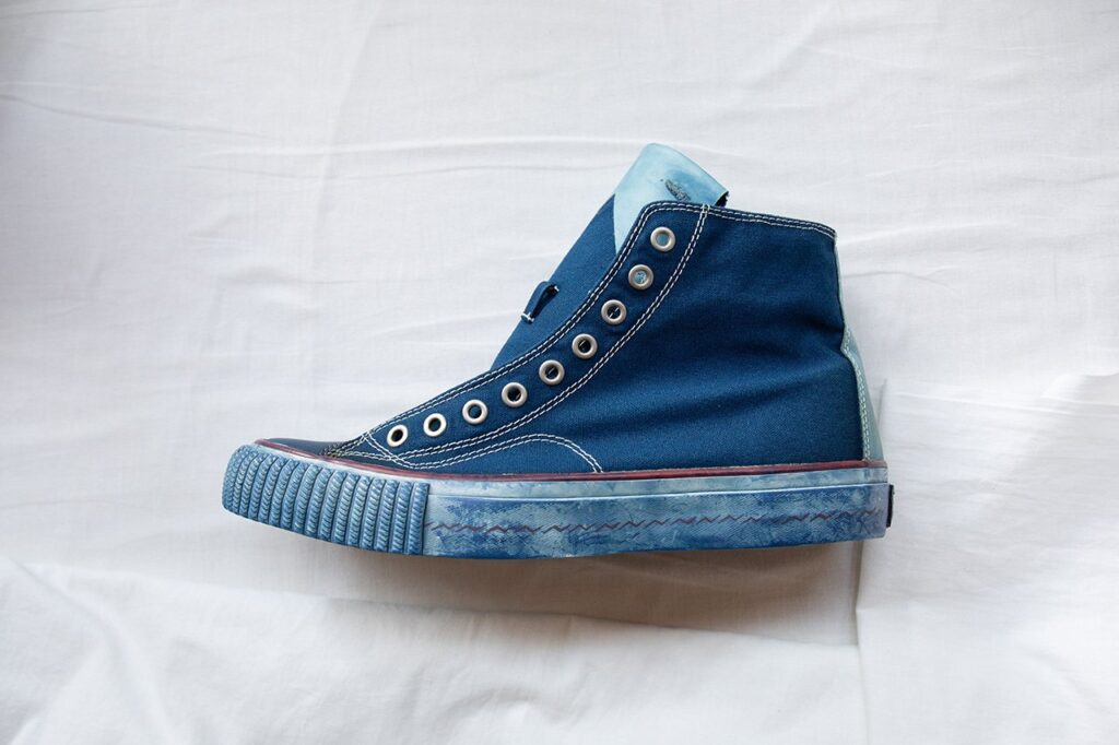 Close-up of an Indigo Camping Trailer high-top shoe hand-dyed in natural indigo, featuring metal eyelets, white contrast stitching, and a textured rubber sole with subtle blue dye variations, showcasing the artisanal craftsmanship and experimental design linked to Visvim’s aesthetic.