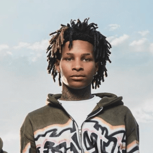 Portrait of rapper Bak Jay wearing a graffiti-patterned zip-up hoodie against a soft blue sky backdrop, embodying the laid-back yet expressive tone of his collaboration with NoCap on “Love on an Island.”