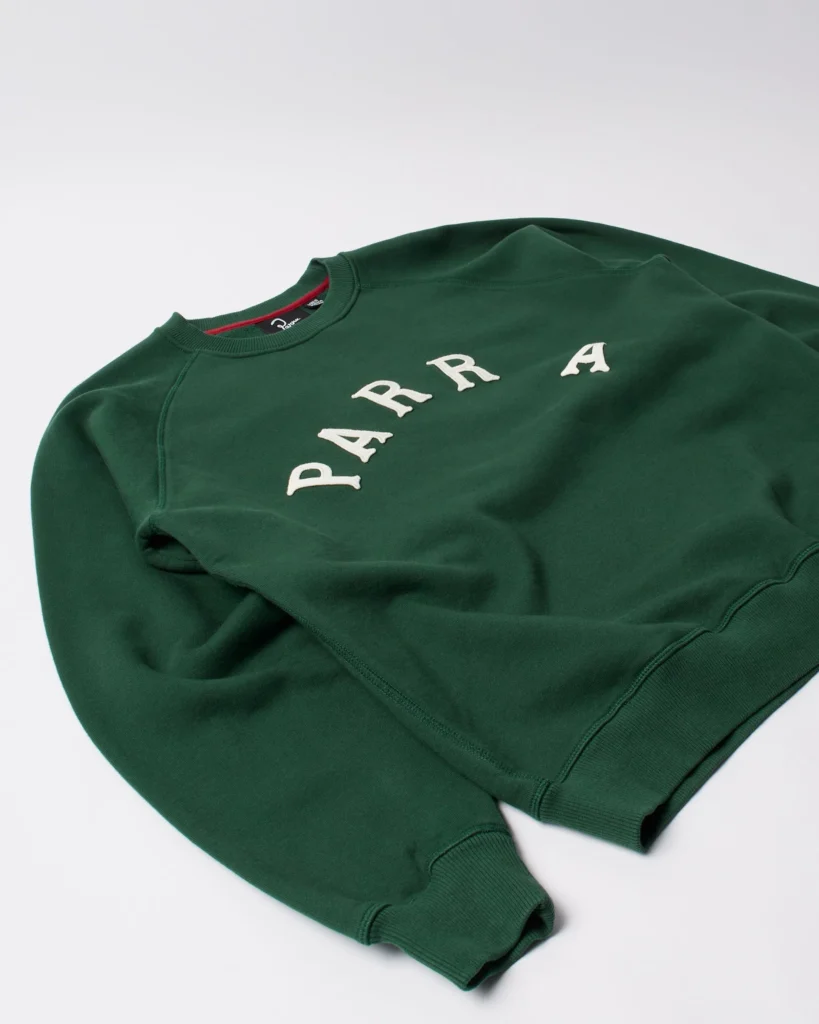 Angled view of the green by Parra crew neck sweatshirt showing the arched cream “PARRA” lettering across the chest, ribbed cuffs, and soft heavyweight cotton texture against a light background.