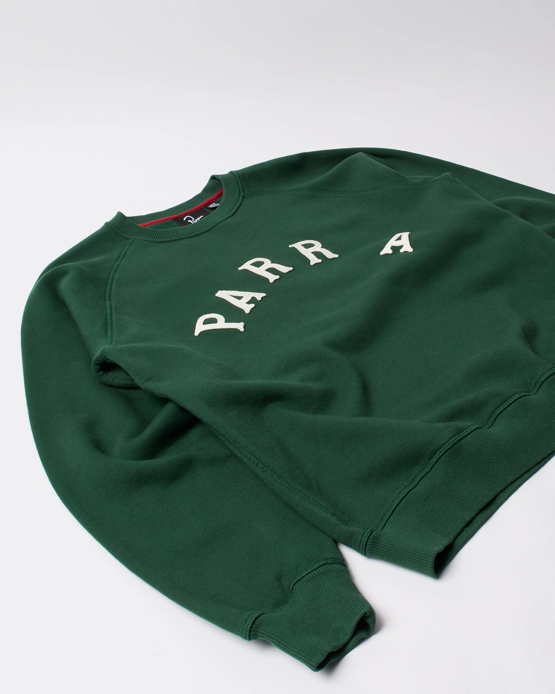 Angled view of the green by Parra crew neck sweatshirt showing the arched cream “PARRA” lettering across the chest, ribbed cuffs, and soft heavyweight cotton texture against a light background.