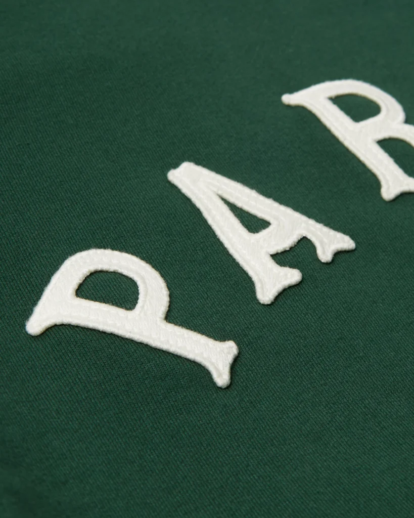Close-up detail of the by Parra crew neck sweatshirt in green, showing the textured cream appliqué “PARRA” lettering stitched onto the heavyweight cotton fabric.