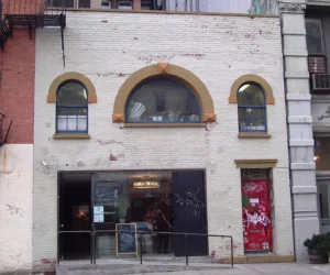 Front façade of 57 Great Jones Street in New York City, the former home and studio of Jean-Michel Basquiat. The two-story brick building features cream-colored paint with weathered patches, arched windows trimmed in ochre, and a distinctive red graffiti-covered door beside a glass-fronted ground-floor entrance.