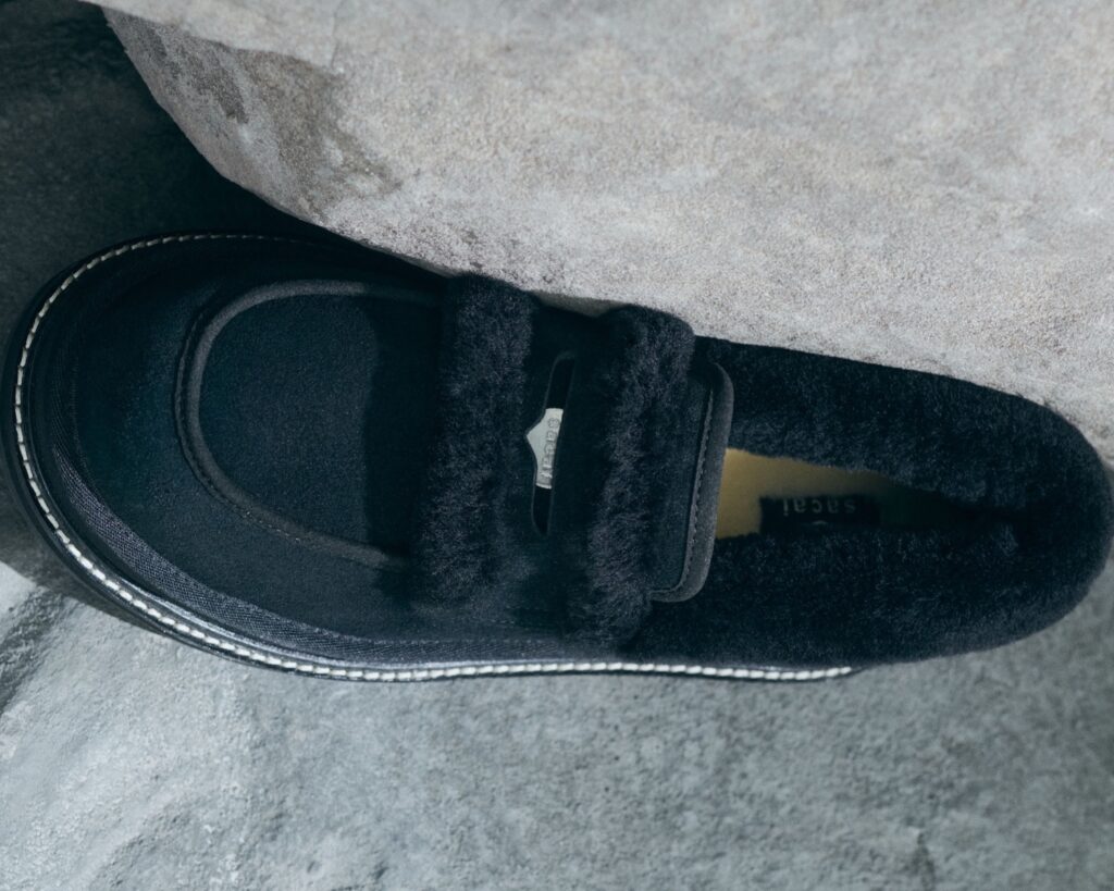 Close-up of the black UGG × sacai loafer from the Autumn/Winter 2025 collection, showcasing its plush shearling lining, suede upper, and stitched detailing that blends luxury craftsmanship with modern comfort.