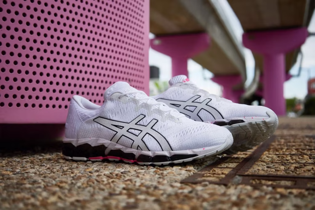 ASICS GEL-QUANTUM 360 5 “Tokyo Neon” shoe photographed in a studio setting. The shoe features a sleek engineered knit upper in black with hot-pink neon highlights across the midsole, heel, and ASICS logo. Transparent GEL cushioning surrounds the midsole, reflecting light like glass. The design draws inspiration from Tokyo’s neon-lit streets, blending performance technology with bold urban style.