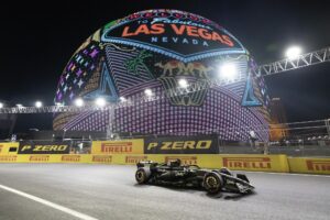 A Formula 1 race car speeds past the illuminated Las Vegas Sphere during the Formula 1 Las Vegas Grand Prix night race, with vibrant neon visuals of the iconic “Welcome to Fabulous Las Vegas” sign glowing overhead.