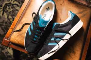 END. x adidas Gazelle Indoor Pro from the “Grass Roots” collection, photographed on textured concrete steps. The shoe features a black mesh upper with suede overlays, teal Three Stripes, and white midsole detailing, symbolizing terrace heritage and END.’s Newcastle roots.