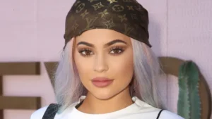 Kylie Jenner Close-up portrait with pastel lavender hair wearing a brown Louis Vuitton headscarf and white top, photographed against a pale pink wall with cactus décor in the background.