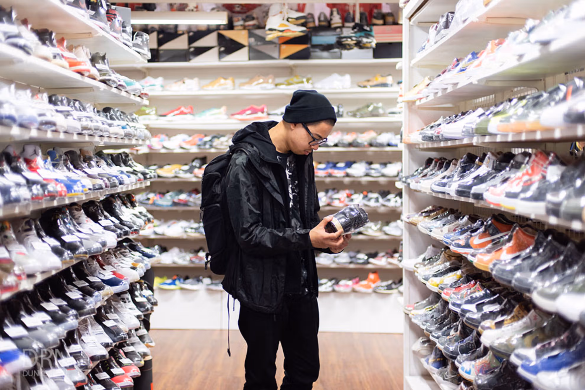 Rolo Tanedo Jr stands in a minimalist sneaker room surrounded by rows of Nike SB Dunks displayed on floating shelves. Each pair—from early HUF Highs to recent collaborations—forms a chromatic timeline of the model’s evolution. Soft studio lighting highlights the suede textures and color-block patterns, reflecting the collector’s meticulous curation and deep connection to skate culture and sneaker history.