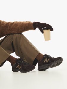 Model wearing the Kith x New Balance 860v2 sneakers in rich brown suede, styled with tailored trousers, brown knitwear, and gloves while holding a coffee cup — showcasing refined street-haute aesthetics.