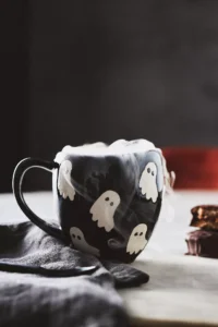 A black ceramic mug decorated with white ghost illustrations, filled with a steaming hot drink, set on a dark cloth napkin with a chocolate treat nearby, evoking a cozy Halloween atmosphere.