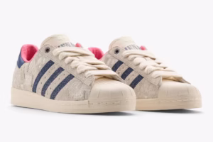 The MENACE x adidas Superstar sneaker featuring a cream suede upper with navy heel tabs, pink satin lining, paisley stitching, and an aged rubber shell toe, photographed on a textured concrete surface highlighting its industrial-inspired design.