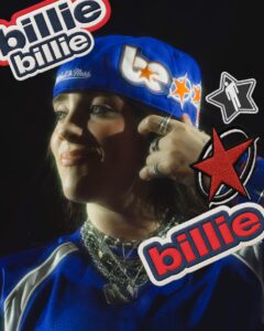 Billie Eilish wearing a blue Mitchell & Ness NBA snapback with her custom ‘BE’ logo and star patches, styled with layered silver necklaces and graphics reading ‘billie’ around the image.