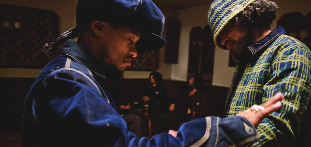 Two men sharing an introspective moment while wearing pieces from the Carhartt WIP × Nicholas Daley Fall/Winter 2025 collection — indigo workwear on the left and tartan herringbone jacket with matching crochet hat on the right, set inside a dimly lit social space evoking reggae-club warmth and community.