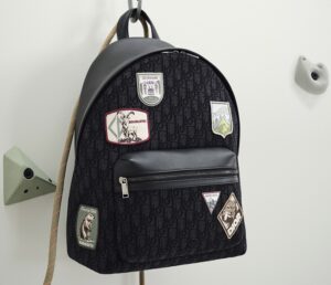 A black Dior backpack from the Spring 2026 Lifestyle Capsule, featuring the brand’s monogram canvas and adorned with embroidered mountain-themed patches. The bag includes a front leather zip pocket and silver hardware, hanging on a minimalist climbing-inspired wall hook—reflecting Dior’s fusion of luxury craftsmanship and alpine adventure.