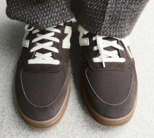 Close-up of the AURALEE x New Balance T500 in Vintage Brown, showing the nubuck and mesh upper with cream laces and signature “N” logo, styled with textured tweed trousers on a light stone surface.