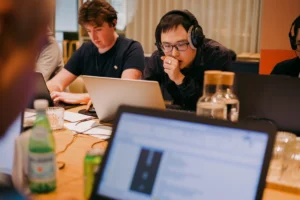 Two focused competitors work on laptops during a live spreadsheet competition; one wears headphones, leaning forward in concentration, while the other types rapidly, with water bottles and soda on the table foreground.