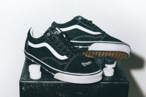 pair of Travis Barker x Vans Old Skool “Dues Paid” sneakers displayed on a dark surface, showing black suede uppers, white leather side stripes, barbed-wire detailing on the midsole, and “Dues” embroidery on the toe box