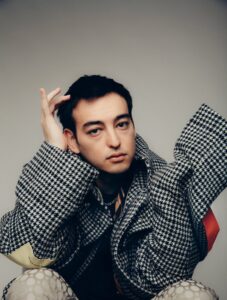 Portrait of a Joji earing a black-and-white houndstooth jacket with oversized sleeves, sitting against a neutral background and gazing thoughtfully at the camera. The styling combines patterned layers and modern textures, evoking a moody, introspective tone consistent