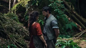 Scene from the 2025 film Hamnet, showing two characters standing close together in a moss-covered forest. The woman, dressed in a red period gown, faces the man wearing a grey linen vest and rolled sleeves, their expressions filled with intimacy and unspoken emotion. The setting evokes nature, love, and the earthy realism central to Chloé Zhao’s direction of the Shakespeare-inspired drama.